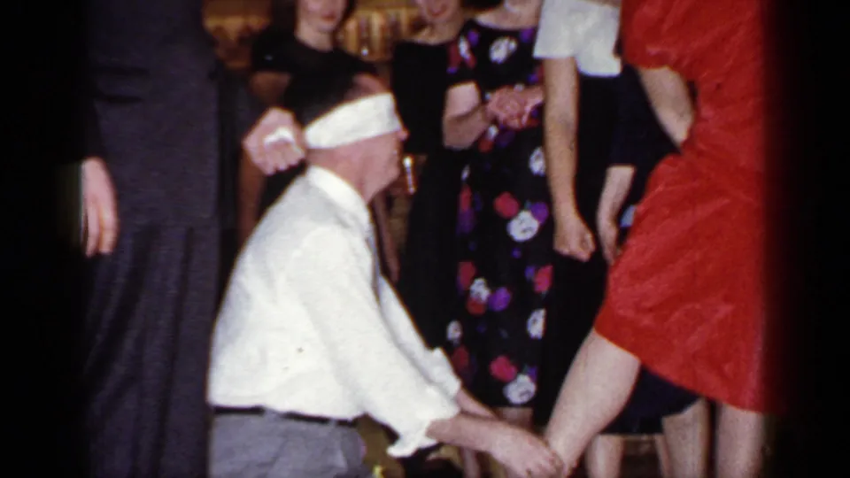 The blindfolded participant remains kneeling during the 1966 party game as the woman in red shifts position in front of guests.