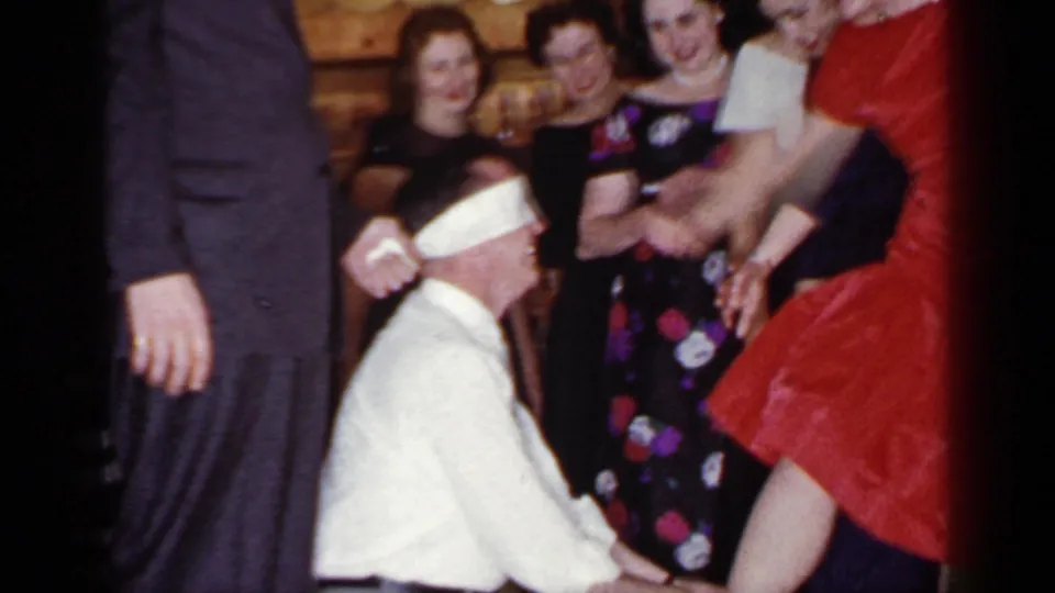 A blindfolded man kneels during the Mineola party game while the woman in red raises a leg and guests smile nearby.