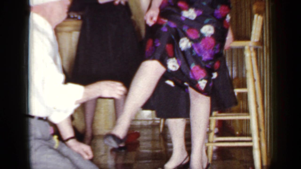 Blindfolded man kneels beside a standing woman in a floral dress during an indoor party game.