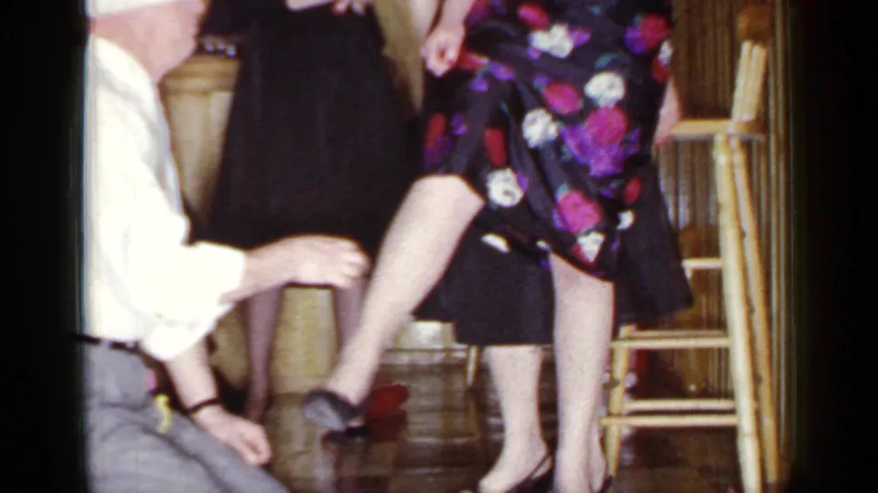 In a 1966 Mineola home-movie frame, a blindfolded man kneels near a woman's leg as adults stand around him indoors.