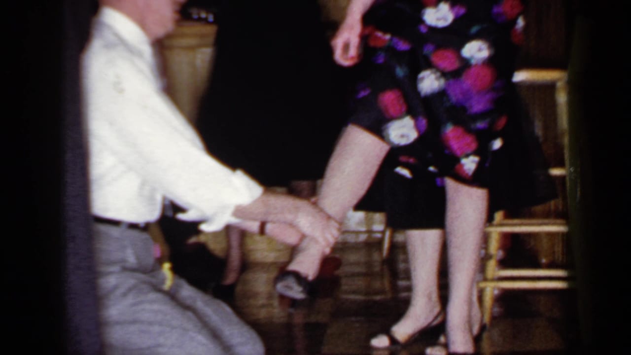 Kneeling blindfolded man holds a woman's ankle while she stands in heels near chairs.