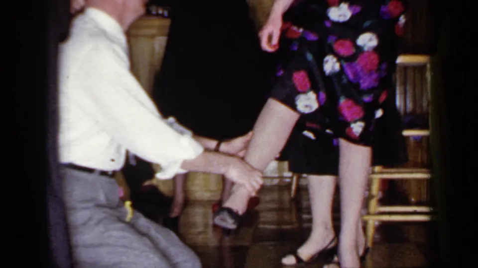 A 1966 home-movie frame shows the kneeling blindfolded man reaching up toward a standing woman's leg in a private interior.
