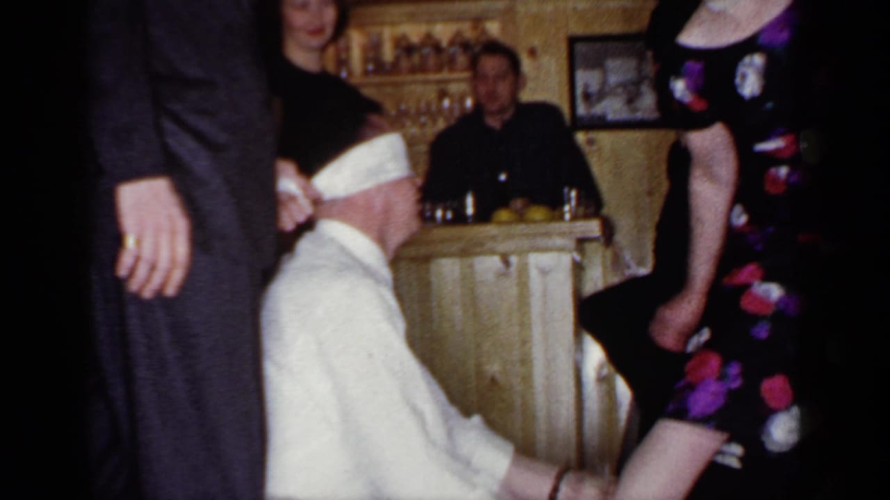 Blindfolded seated man reaches toward guests in a wood-paneled party room.