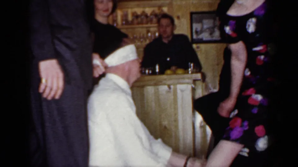 In the 1966 Mineola home-movie footage, a blindfolded seated man reaches toward standing guests near a home bar.