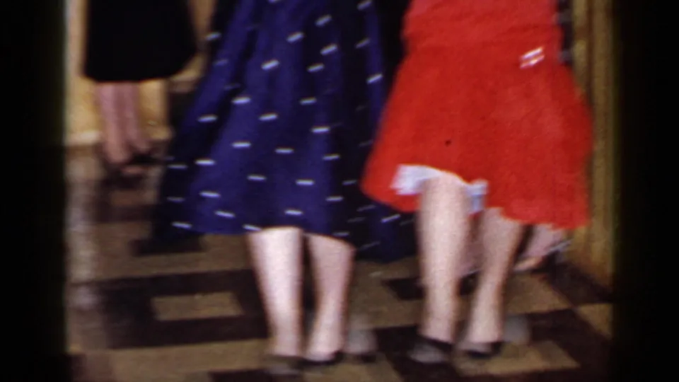 Two women in colorful dresses dance in the Mineola party interior while other adults stand nearby.
