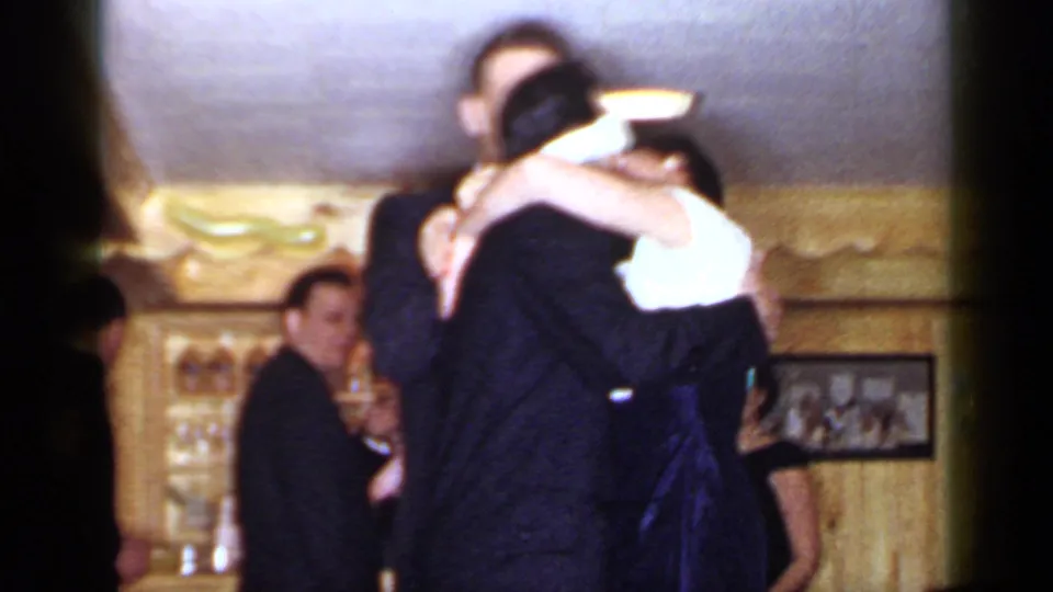 1966 Mineola home-movie frame of a suited man embracing a woman in a white top while other adults stand in a paneled interior.