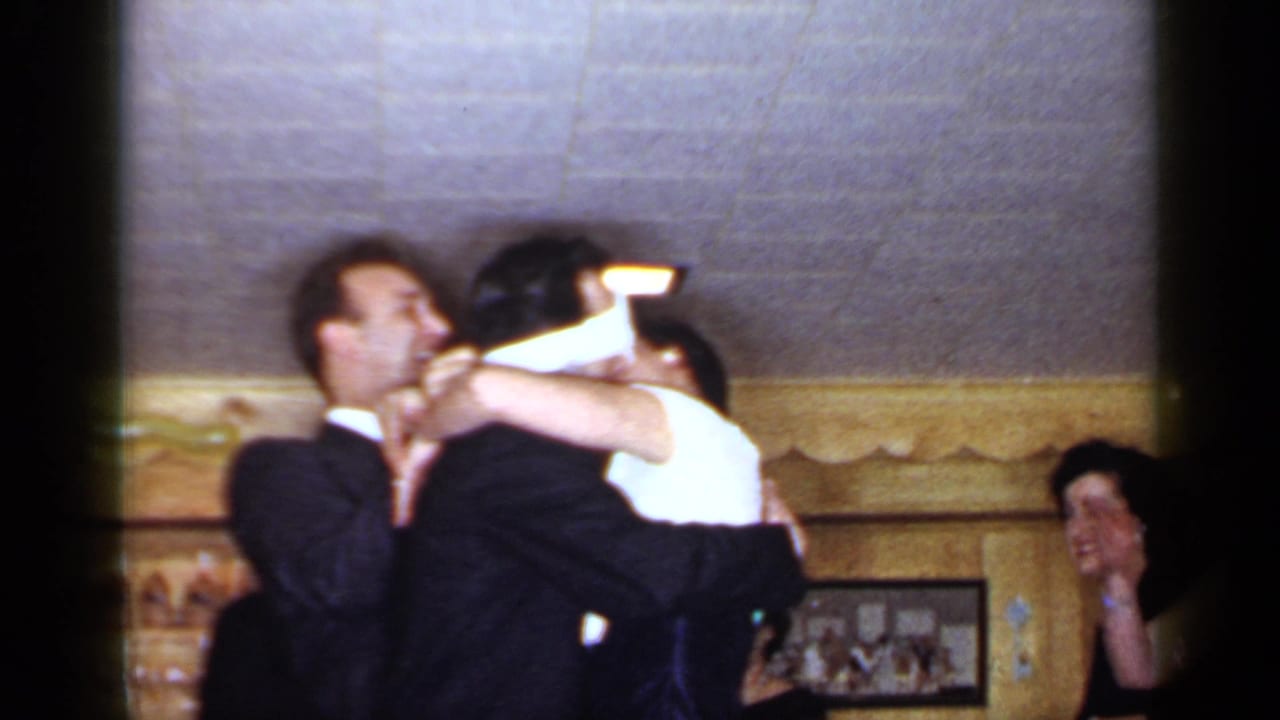 Man in a dark suit embraces a blindfolded woman as guests watch indoors.