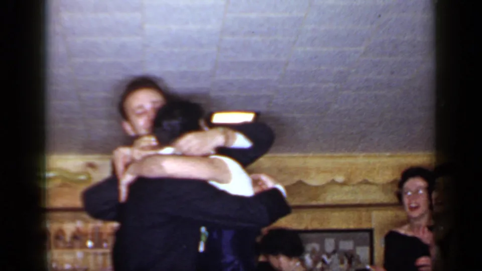 1966 Mineola wedding reception frame with the blindfolded pair embracing closely and a laughing guest visible behind them.