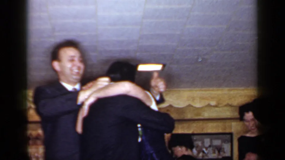 1966 Mineola reception frame showing a smiling suited man and blindfolded woman embracing before guests in a private interior.