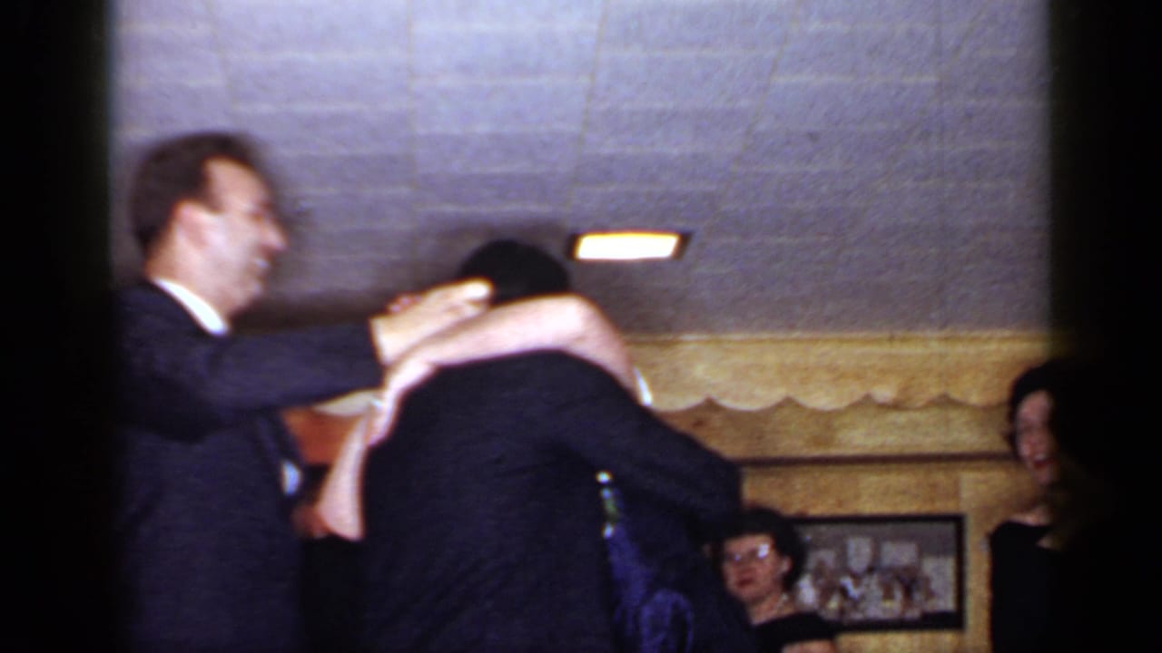 Two men in dark suits lean into an embrace while people watch in a wood-paneled room.