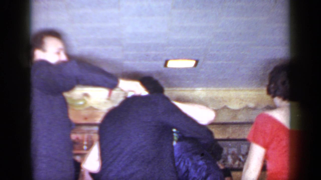 1966 Mineola frame with a suited man reaching around another person at a formal indoor gathering as a woman in red stands nearby.
