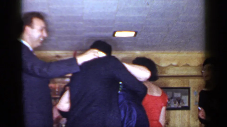 1966 Mineola home-movie still showing adults gathered closely in a wood-paneled interior, with a suited man smiling and a woman in red nearby.