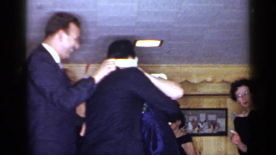 1966 Mineola frame showing a smiling suited man beside another guest during an indoor social gathering, with women watching in the background.