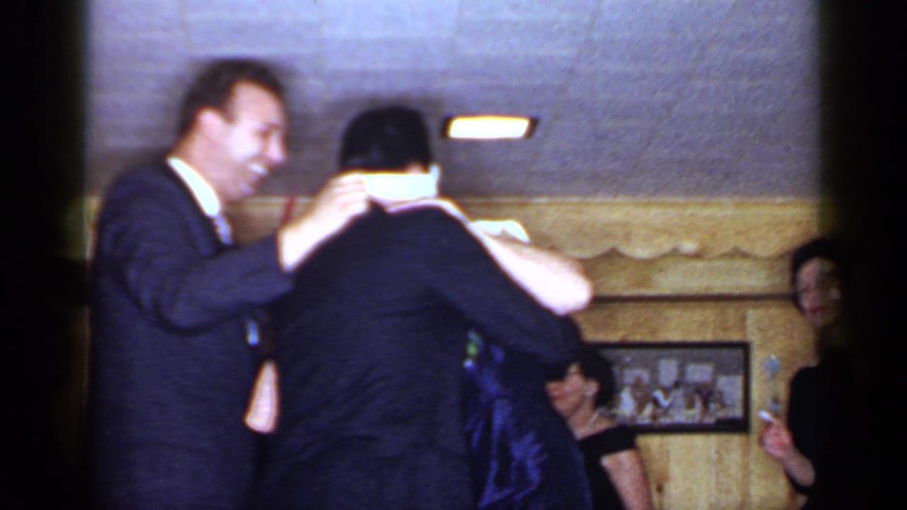 Adults at a party as a man helps blindfold another guest in a paneled room.