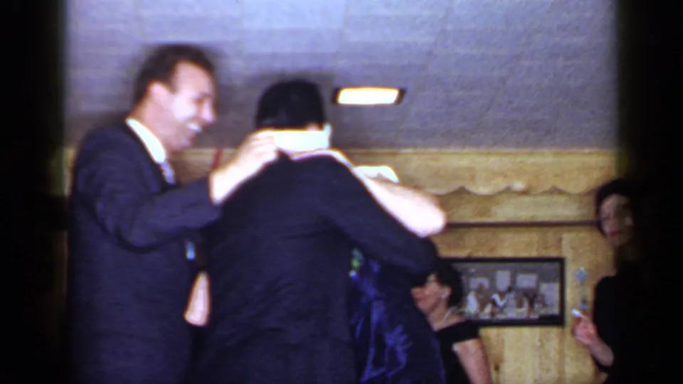 1966 Mineola home-movie frame showing adults in formal clothing during a blindfold party game in a wood-paneled interior.