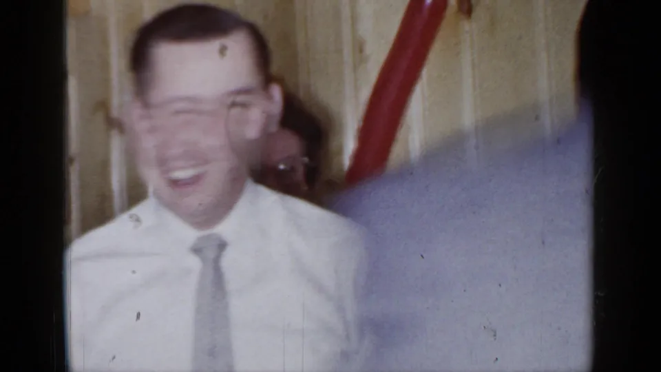 A smiling adult man in a white shirt and tie reacts during the 1966 Mineola party game in a private interior.