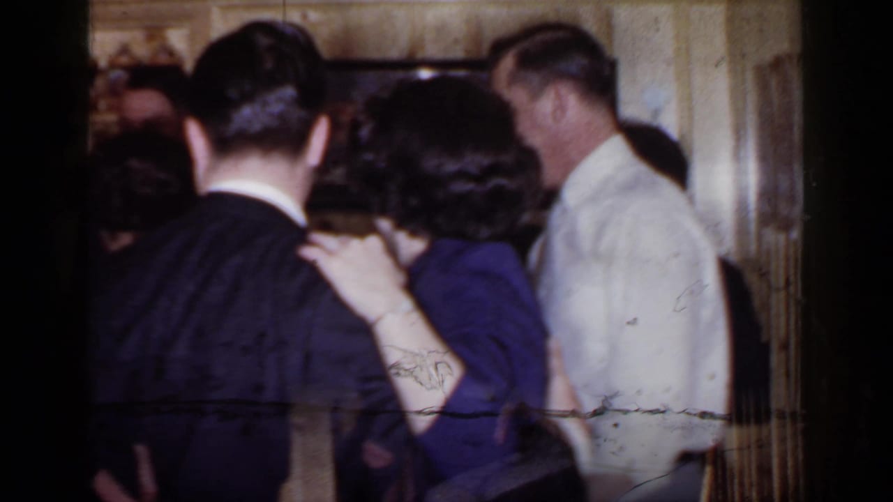 Adults in suits and dresses dance closely in a wood-paneled party room.