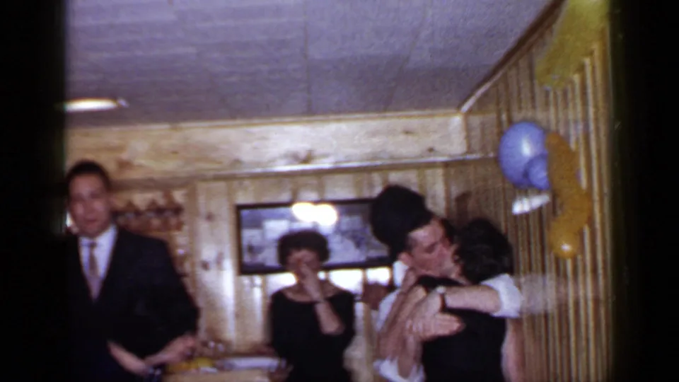 1966 Mineola home-movie frame showing a suited guest at left and a couple kissing near balloons in a wood-paneled room.