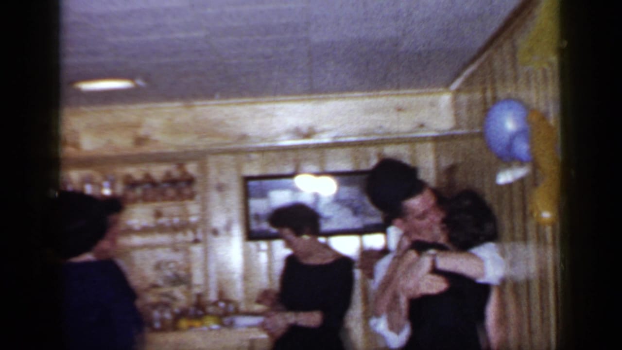 Adults in formal clothing gather at a wood-paneled bar while a couple embraces near balloons.