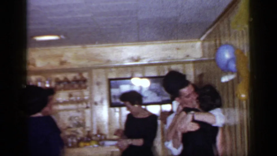 1966 Mineola home-movie frame showing reunion guests in suits and dresses around a wood-paneled bar as a couple embraces.