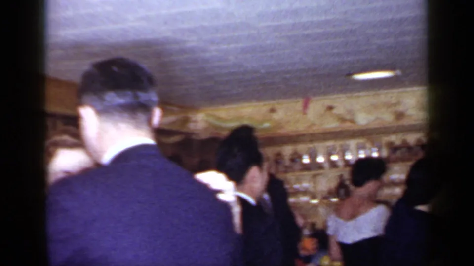 Handheld 1966 Mineola reunion frame showing adults packed into a dim wood-paneled bar, with suits, dresses, and drinks visible.