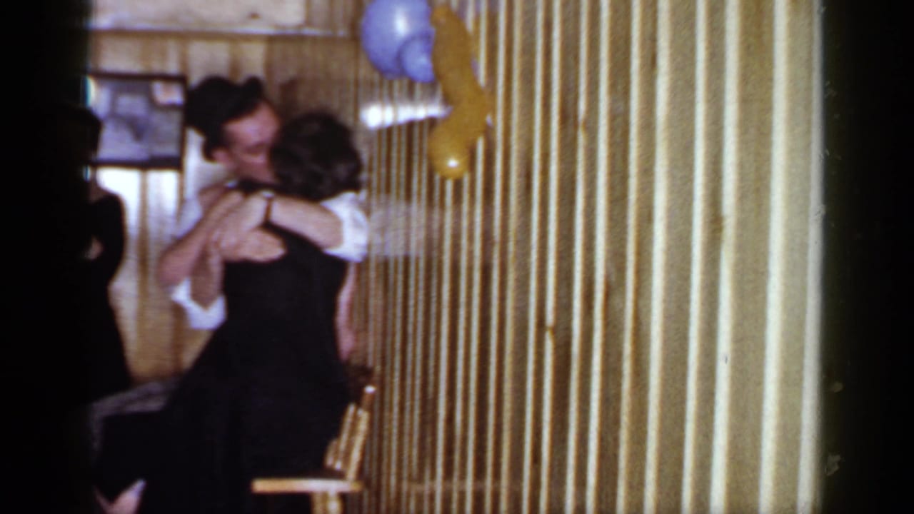 Adults in formal clothing embrace or dance beside balloons in a wood-paneled room.