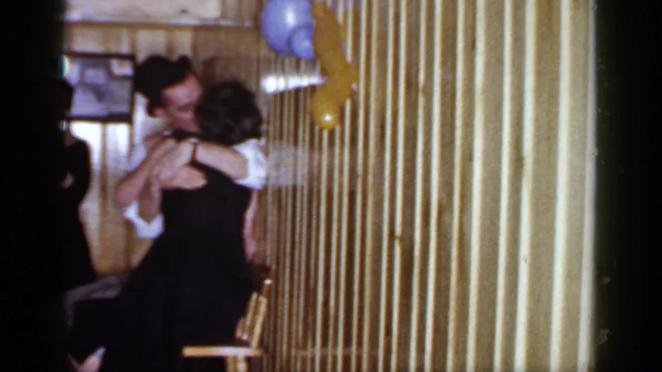In the 1966 Mineola home-movie frame, formally dressed adults dance or embrace near balloons and chairs in a private wood-paneled interior.