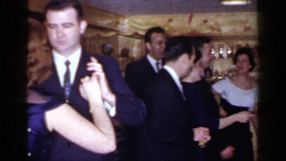 A suited man dances in the foreground while other formally dressed adults socialize in the 1966 Mineola party interior.