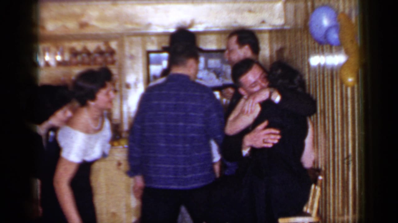 Adults gathered at a wooden bar while a couple hugs near the right side.