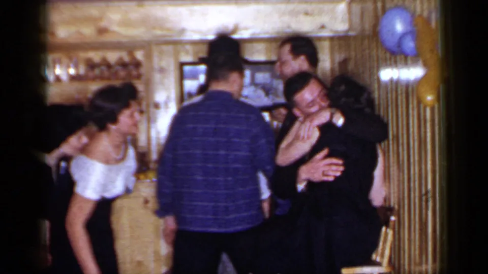 1966 Mineola home-movie frame showing adults at a wood-paneled bar, with a woman in a white dress and a couple embracing nearby.