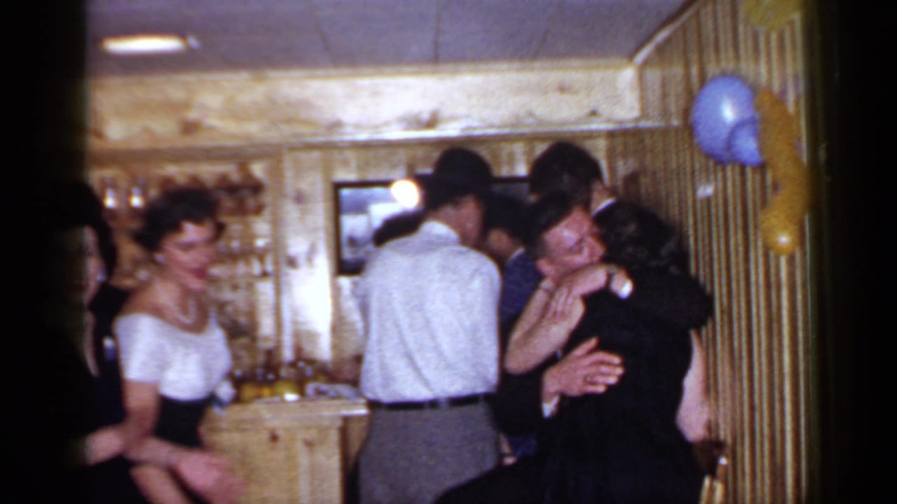 Party guests stand at a wooden bar, with a couple embracing at right.