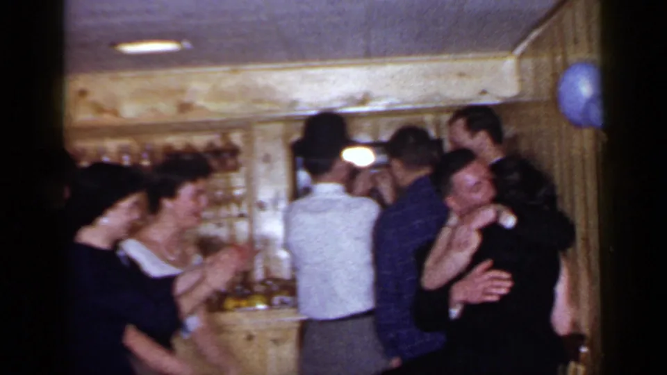 1966 Mineola home-movie frame showing adults clapping and talking in a crowded wood-paneled bar interior with bottles behind them.