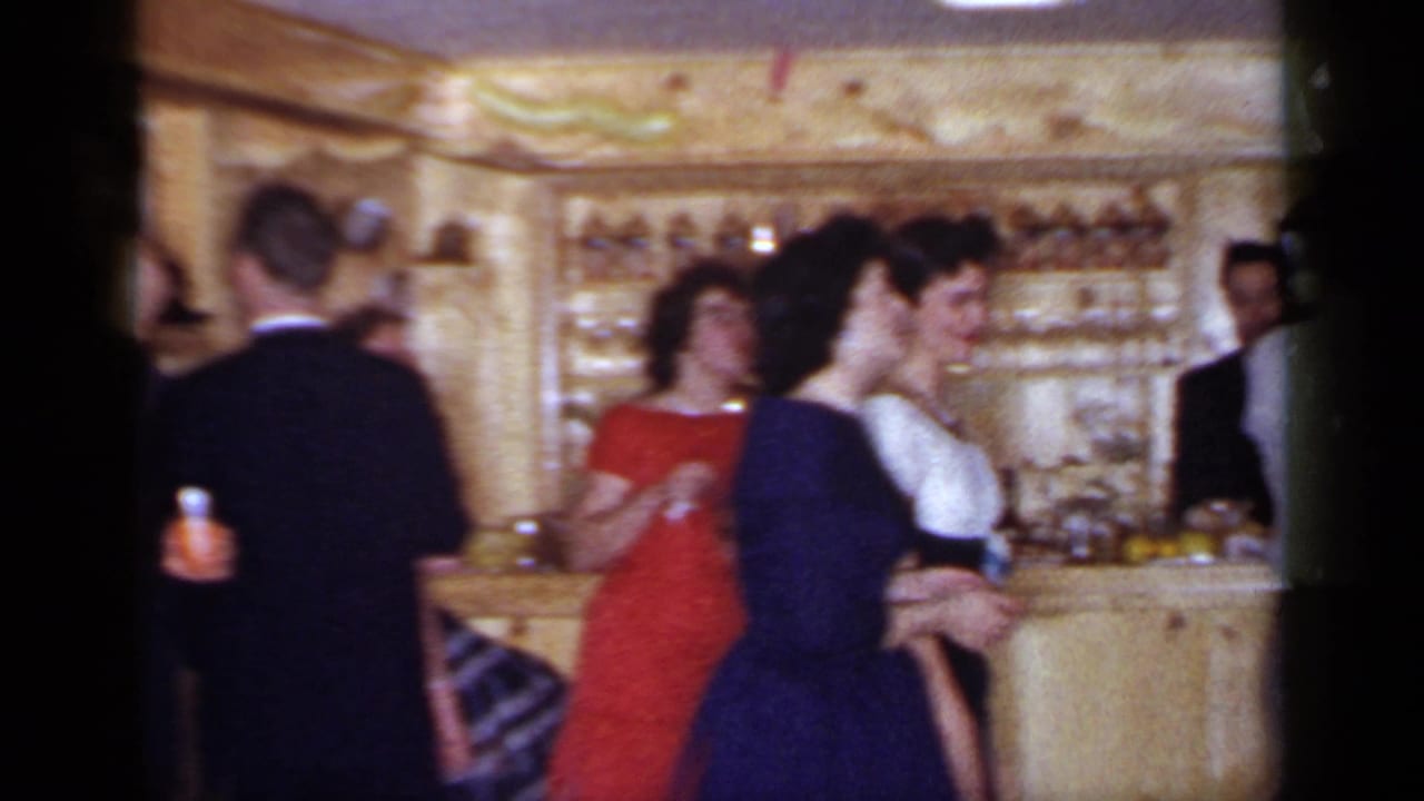 Adults in formal clothing stand near a wood-paneled bar during a party.