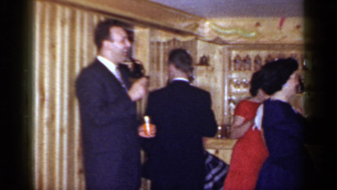 1966 Mineola party footage with a suited man holding a drink as guests mingle in a bar interior.