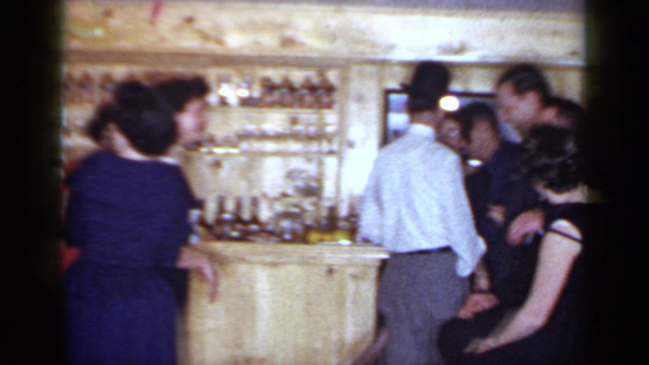1966 Mineola home-movie frame of adults gathered around a wood-paneled bar during a social event.