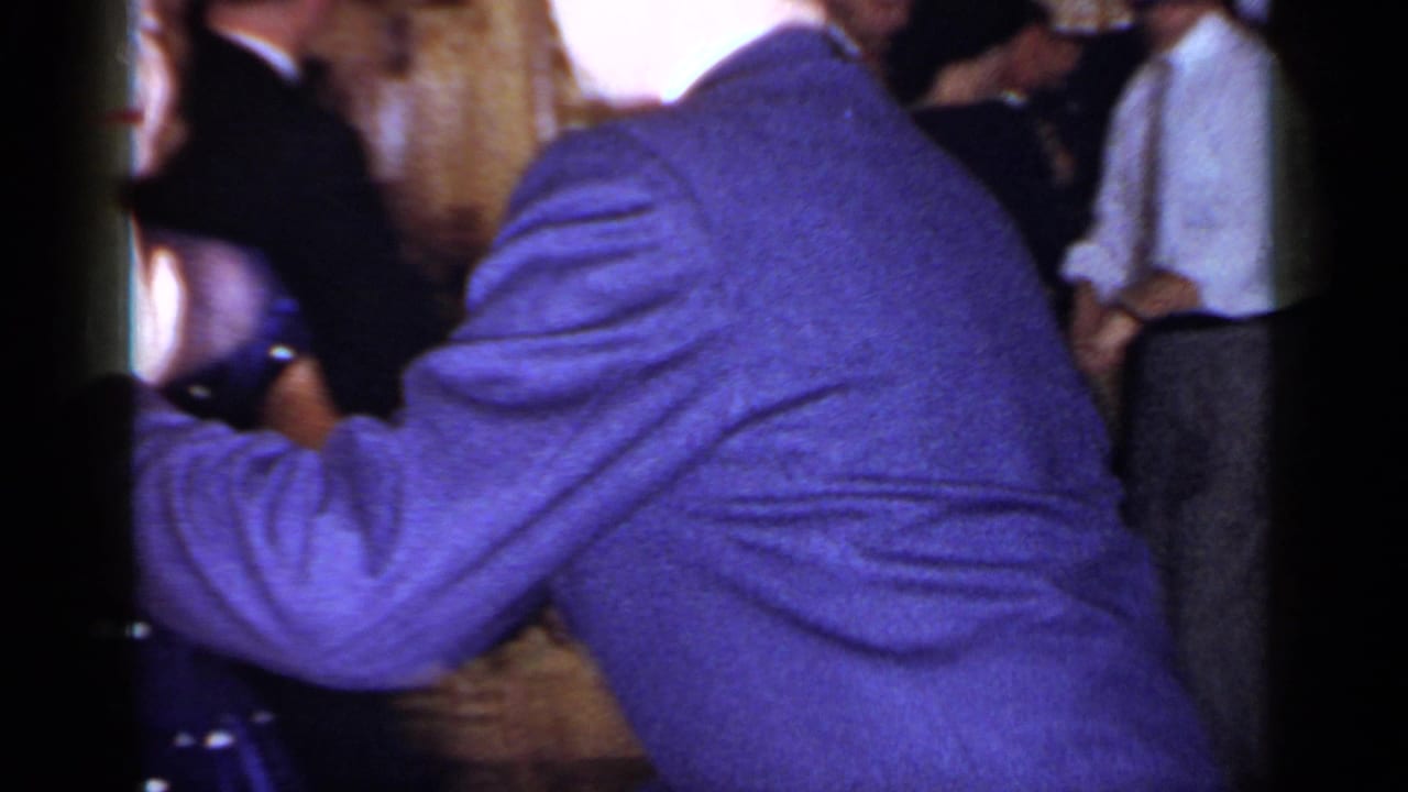 A man in a dark suit dances closely with a woman in a blue patterned dress indoors.