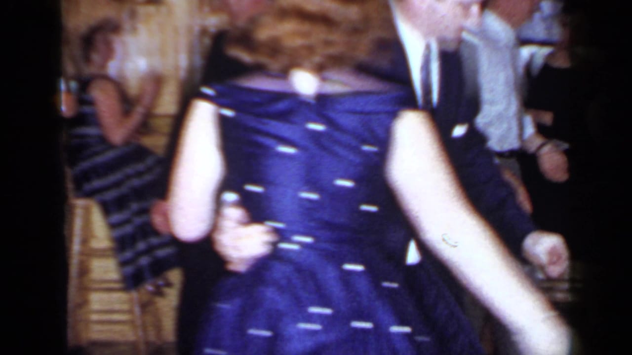 Adults dancing in a crowded interior, including a woman in a blue polka-dot dress.