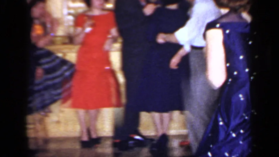 1966 Mineola home-movie frame with adults dancing indoors, including a red dress, dark dresses, and a blue polka-dot dress at right.