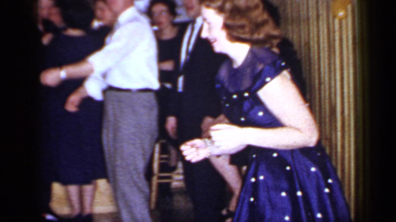 Adults dancing at an indoor party while a woman in a dark polka-dot dress moves in the foreground.