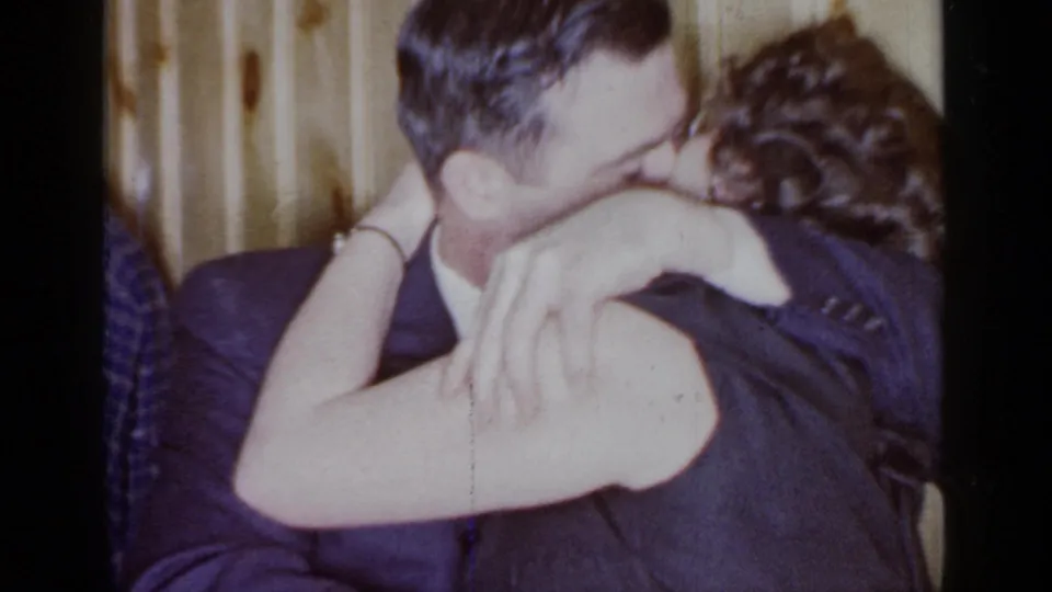 1966 Mineola home-movie frame of an adult couple kissing, with a partial guest figure and paneled interior behind them.