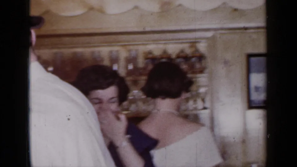 Women in evening dresses stand near bar shelves during the 1966 Mineola indoor party footage.