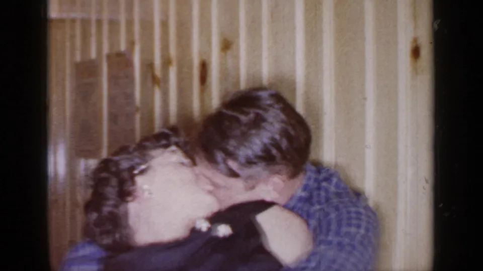 A couple shares a close kiss inside a wood-paneled Mineola interior in 1966 home-movie footage.
