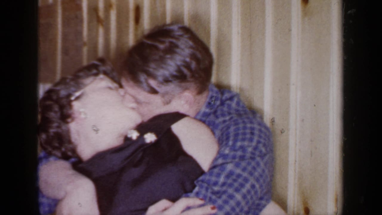 Couple embracing and kissing in front of wood-paneled walls.