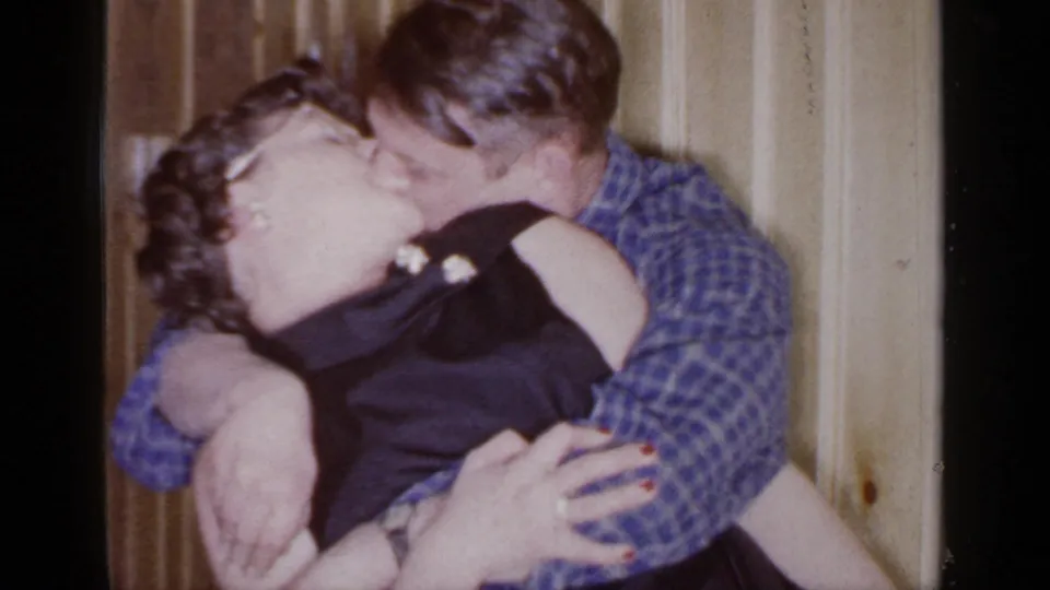 The couple continues kissing in the Mineola interior, framed close against vertical wooden paneling.
