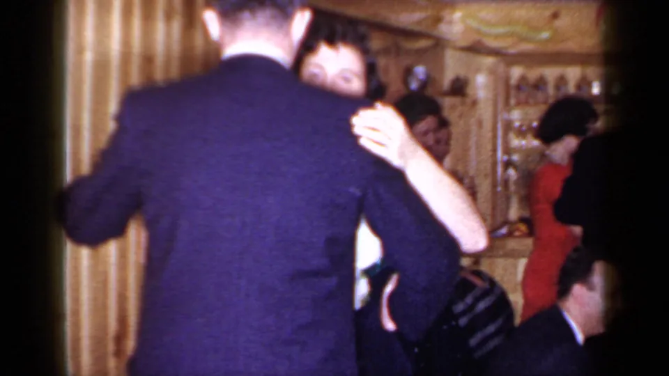The dancing couple moves through the Mineola interior as other adults sit and stand around the wood-paneled room.