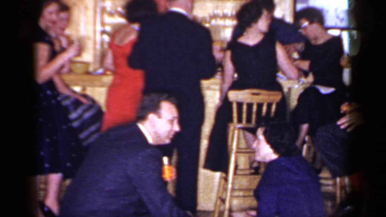 Adults stand and sit around a bar, with drinks and stools visible.