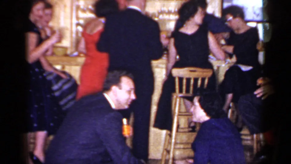A group of adults in formal evening clothing gathers around a wooden bar in the 1966 Mineola home-movie scene.
