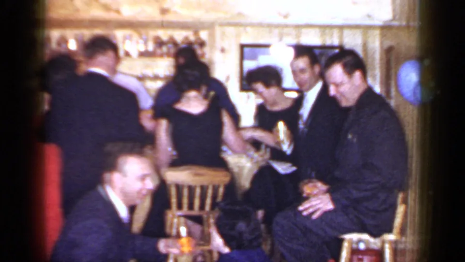 Adults in suits and dresses socialize around a wooden bar in Mineola, New York, from the 1966 8mm home-movie clip.