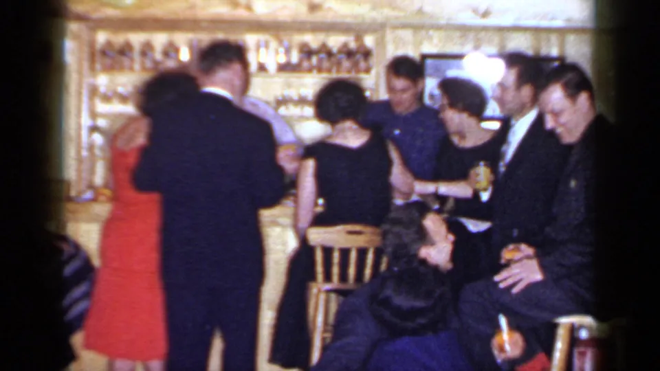 A barroom group talks and drinks while a couple stands together in this 1966 Mineola, New York home-movie frame.