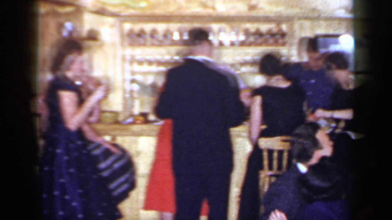 Adults in formal clothing gather near a bar while a couple dances in a warm interior.